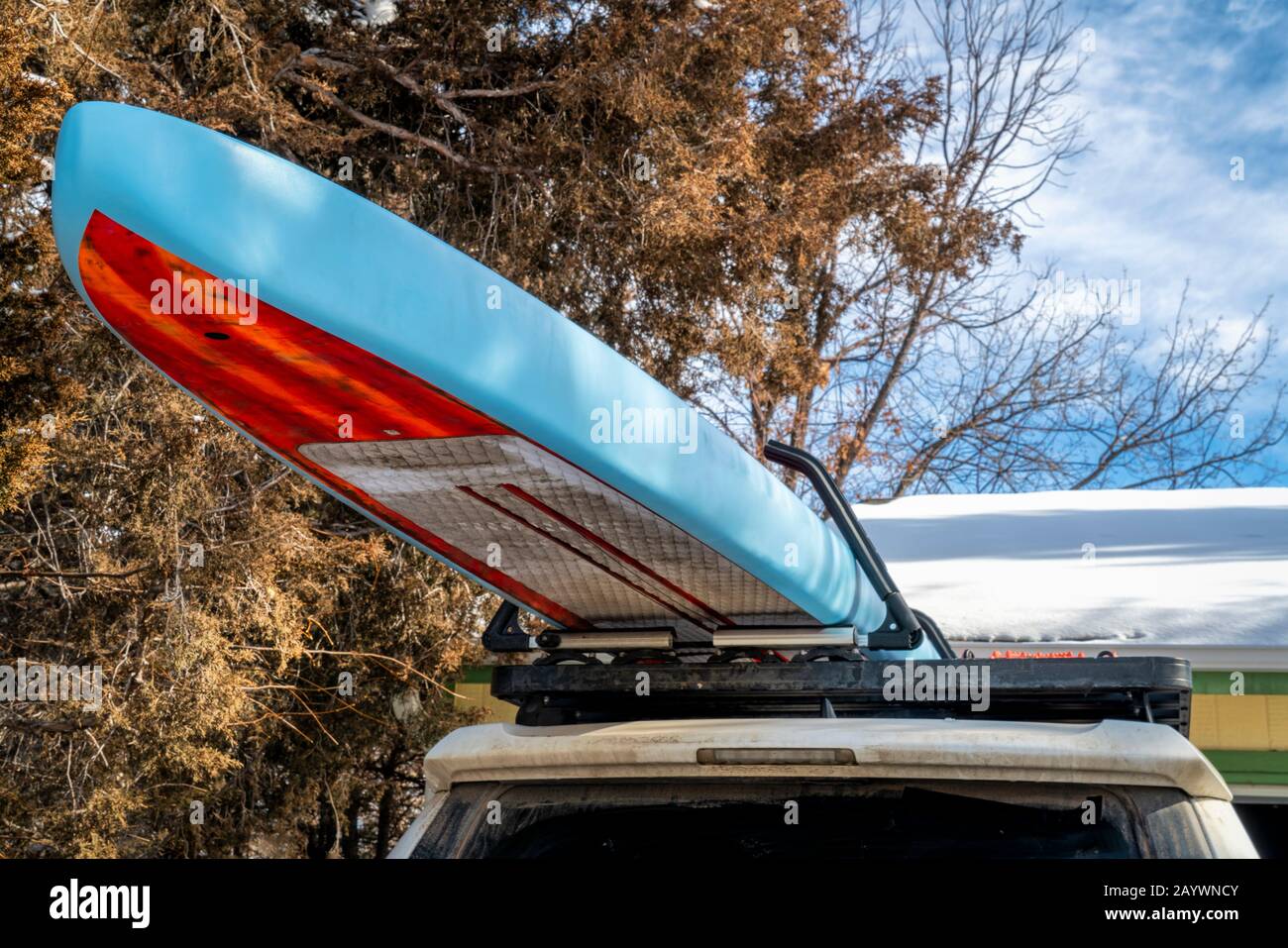 Unbegrenztes Rennfahrer-Stehpaddleboard auf den Dachregalen des SUV-Autos in einer Einfahrt, sowohl schmutzig als auch schlammig nach langer Winterreise. Stockfoto