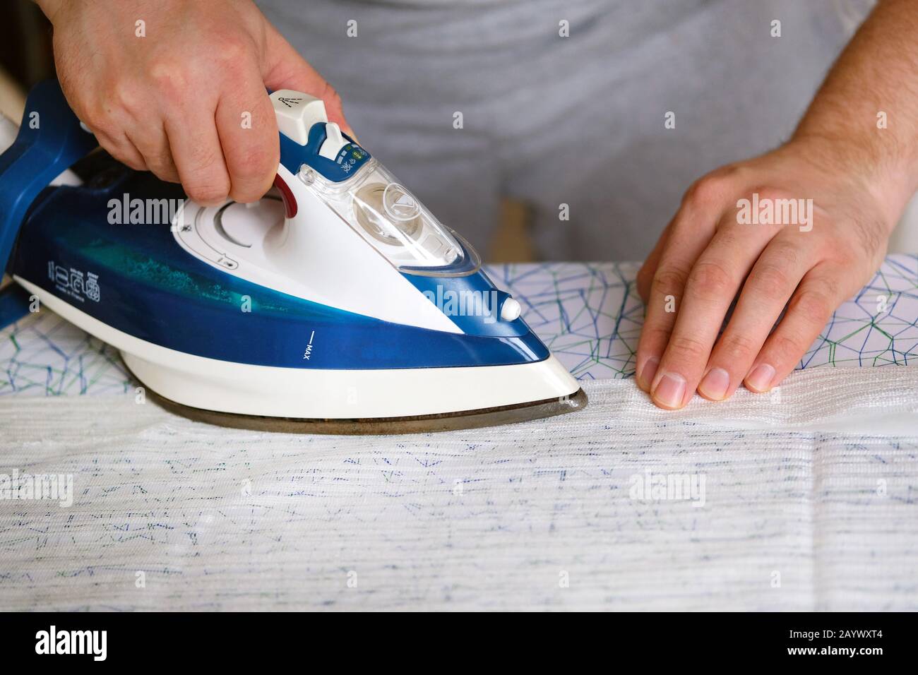 Hausarbeit und Haushaltskonzept. Mann bügelt zu Hause Textil auf dem Bügelbrett. Blaues Eisen in der Hand des Menschen. Stockfoto Hausarbeit und Haushaltskonzept. Mann bügelt zu Hause Textil auf dem Bügelbrett. Blaues Eisen in der Hand des Menschen. Stockfoto