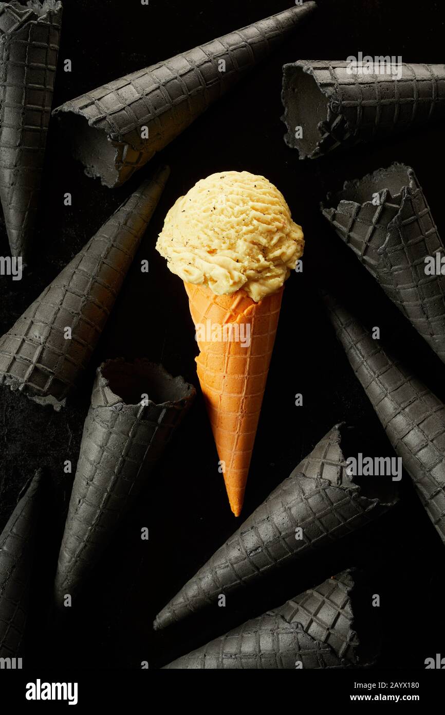 Einfarbige gelbe Eisschaufel in einem Kegel zwischen verstreuten leeren monochromatischen grauen Kornets über schwarzem Hintergrund in einer konzeptuellen ima-Lebensmittel Stockfoto