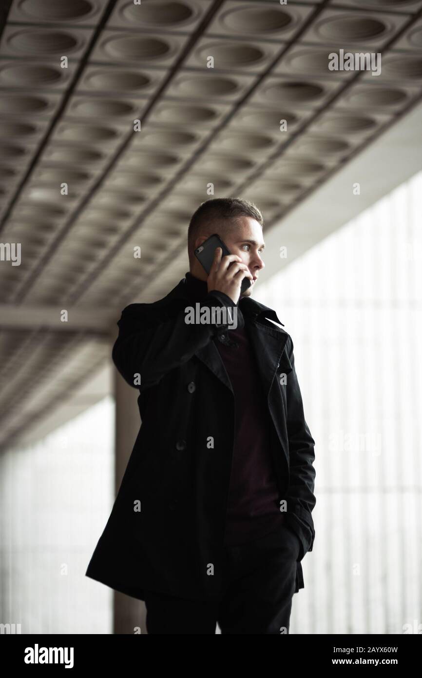 Stilvoller Mann in schwarzem Trenchcoat und Polo-Gespräch auf einem Handy Stockfoto