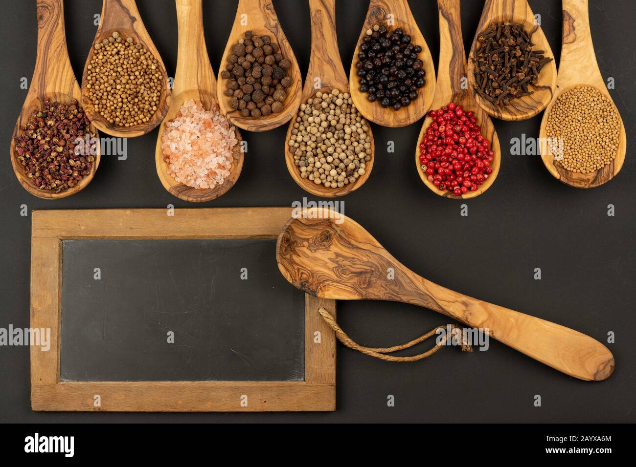 Gewürz und Essen - Neun Kochlöffel aus olivem Holz in einer Reihe mit Gewürz- und Pfeffersorten und einem alten schwarzen Schiefertafel mit Kopierraum Stockfoto