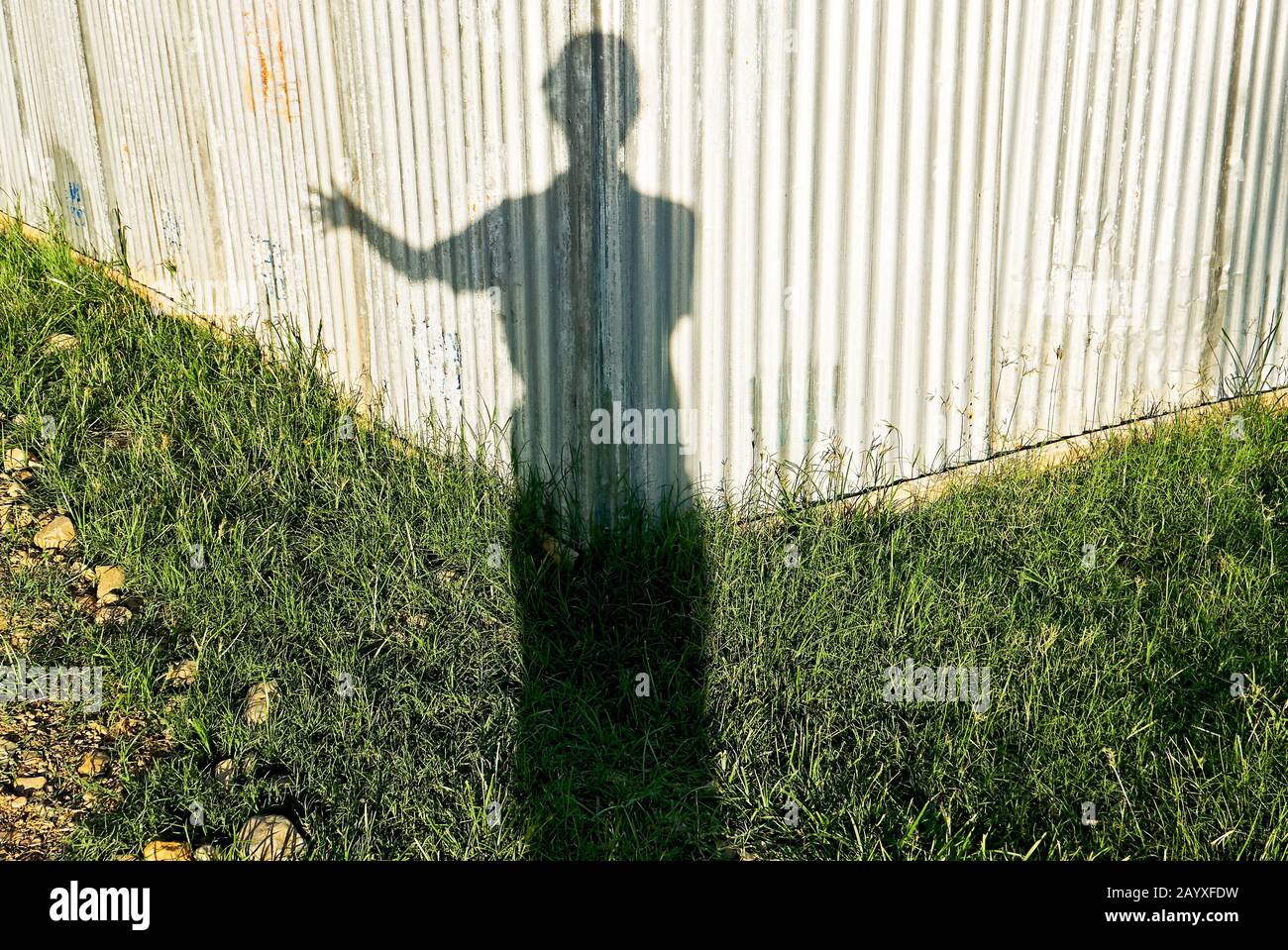 Ein Schatten eines jungen männlichen Menschen, der nur mit einer Hand grüßt und vor einer sonnenbeleuchteten Wellblechwand und einer Rasenfläche steht Stockfoto