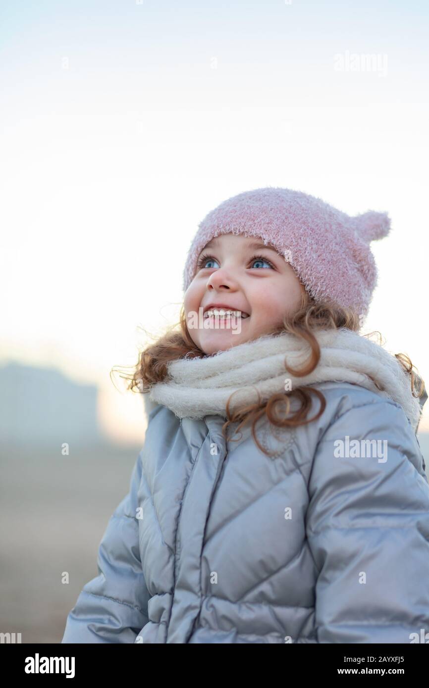 Das entzückende kleine Mädchen lächelt und blickt auf den Himmel. Ausdruck der Freude mit blauen Augen und blonden Haaren, Winterkleidung. Stockfoto