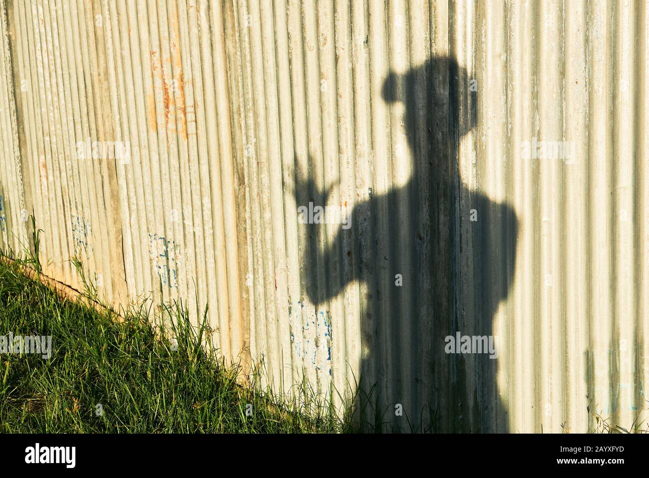 Ein Schatten eines jungen männlichen Menschen mit Hut, nur mit einer Hand grüßenden Gruß, der bei Sonnenuntergang vor einer Wellblechwand und einer Rasenfläche steht Stockfoto
