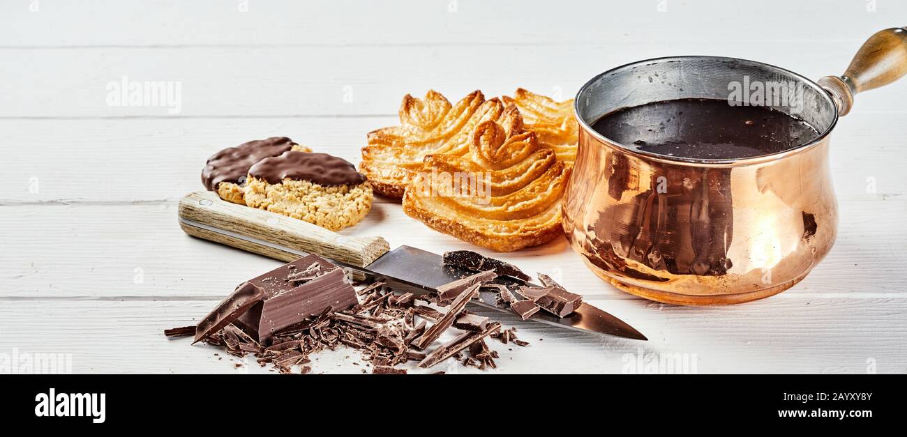 Frisch gebackenes Gebäck in geschmolzener Schokolade in einem glänzenden Kupfertopf mit geschachtelter Süßigkeit und einem Messer im Vordergrund über weißem Holz in einer Pfanne Stockfoto