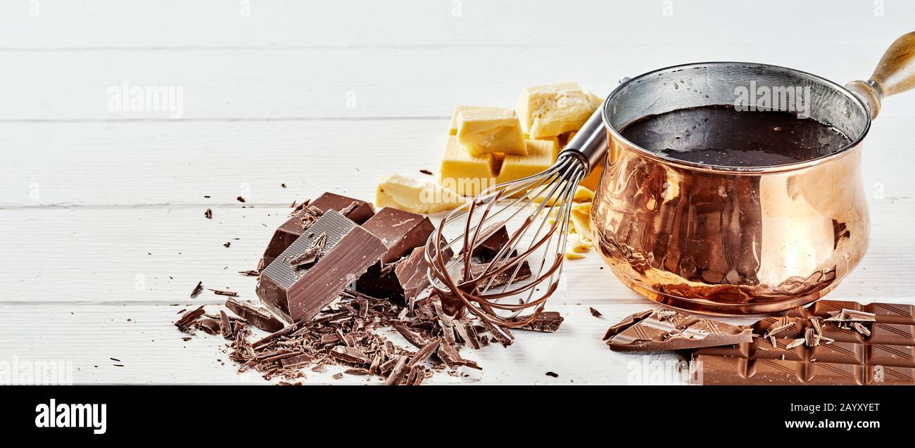 Aufschlagen einer Soße geschmolzener Schokolade in einer Kupferpfanne mit einem Handschaufelchen und verschiedenen Stäben aus gekopffter Schokolade über einem weißen Holzhintergrund mit Kopie Stockfoto