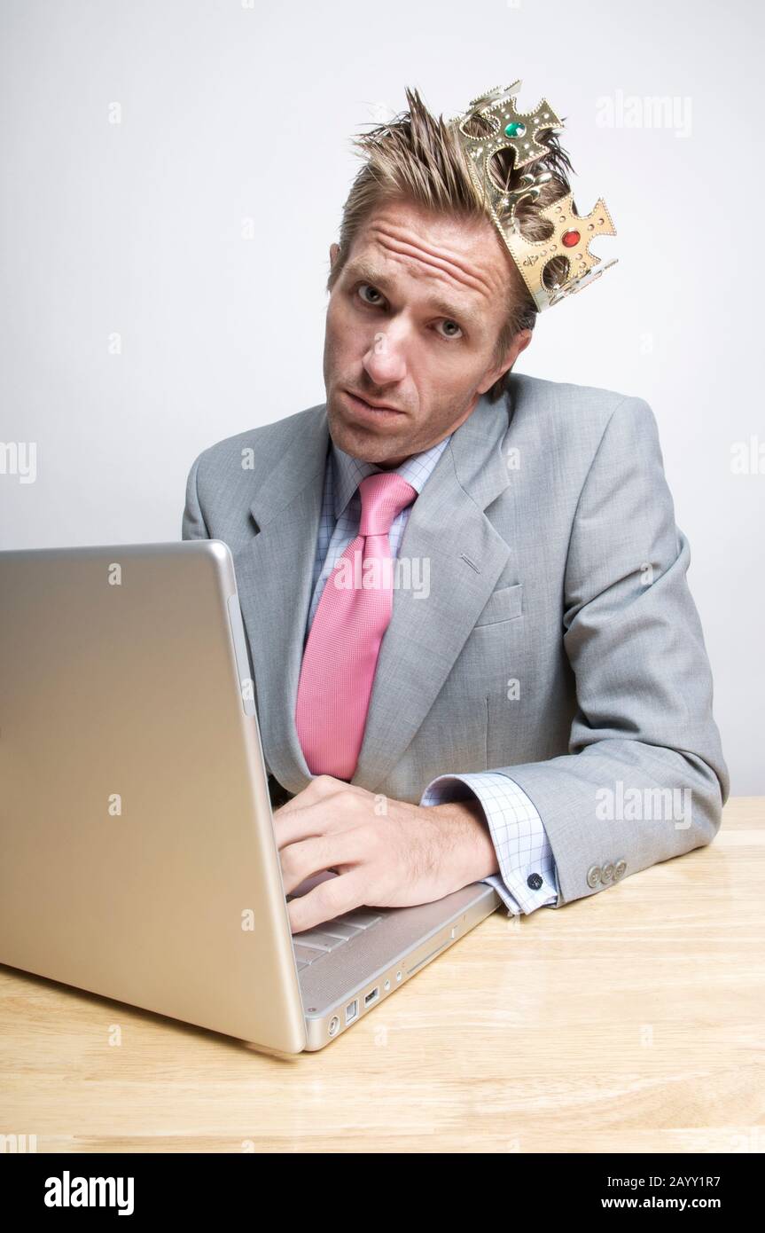 Büroangestellter mit goldener Krone an seinem Schreibtisch mit einem selbstwichtigen Ausdruck sitzend Stockfoto