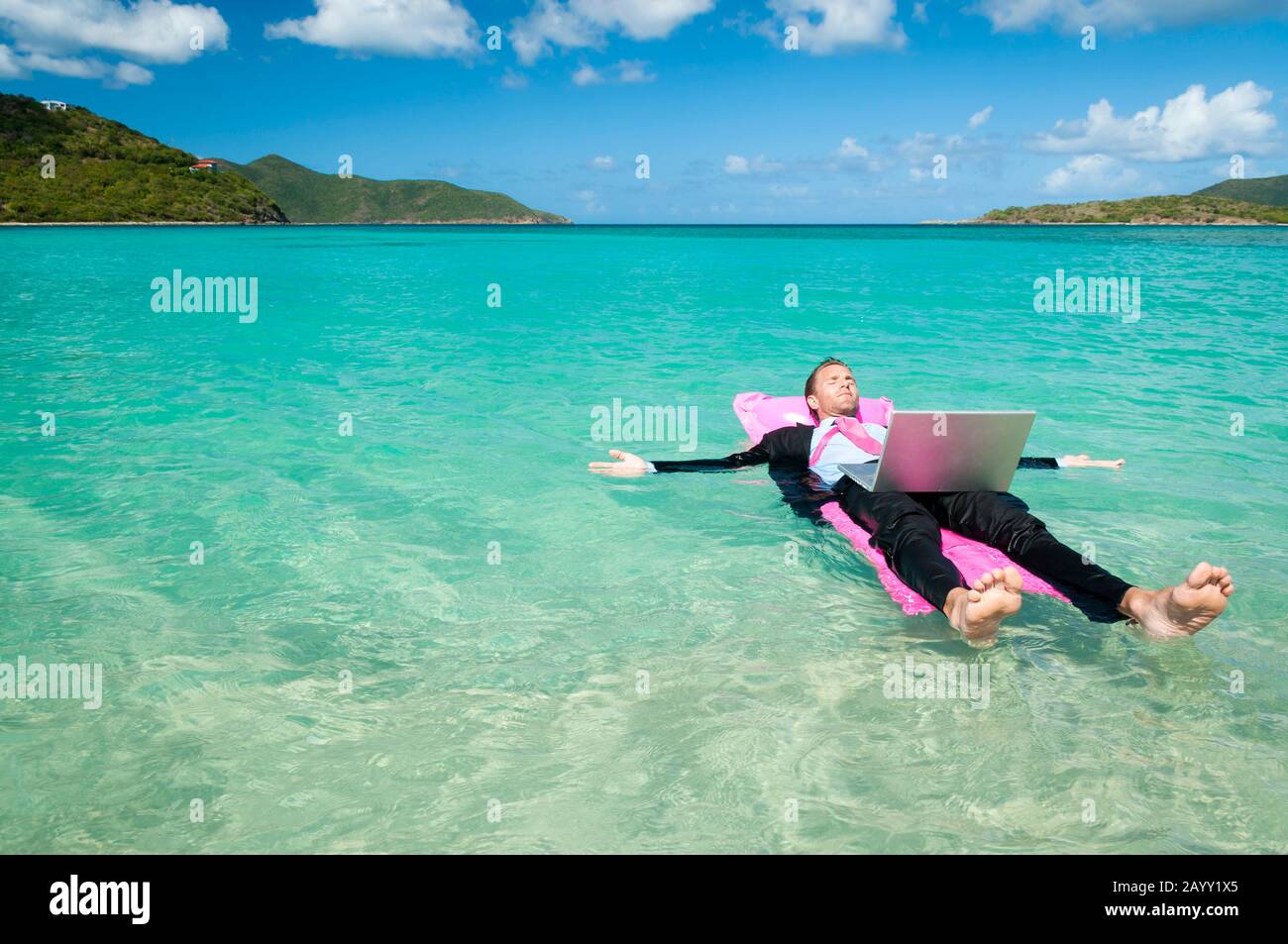 Büroangestellte, die mit seinem Laptop auf einem pinkfarbenen, aufblasbaren Floß im hellen tropischen Meer schwimmen Stockfoto