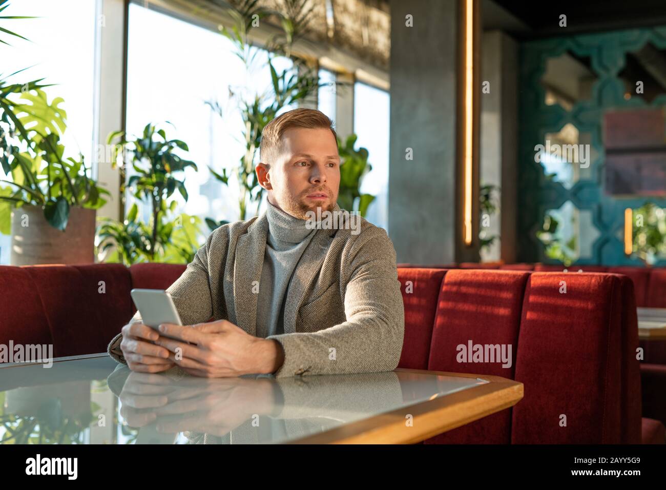 Horizontales Portrait des jungen erwachsenen kaukasischen Mannes, der in einem modernen Café sitzt, in dem das Smartphone auf jemanden wartet Stockfoto