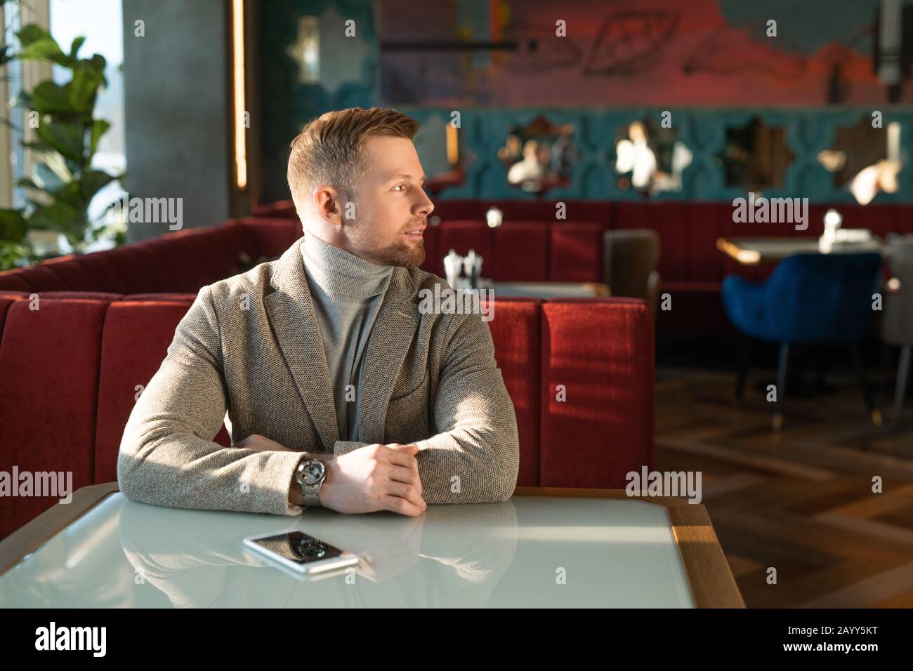Horizontales Porträt eines jungen erwachsenen kaukasischen Mannes, der allein am Café-Tisch sitzt und auf jemanden wartet Stockfoto