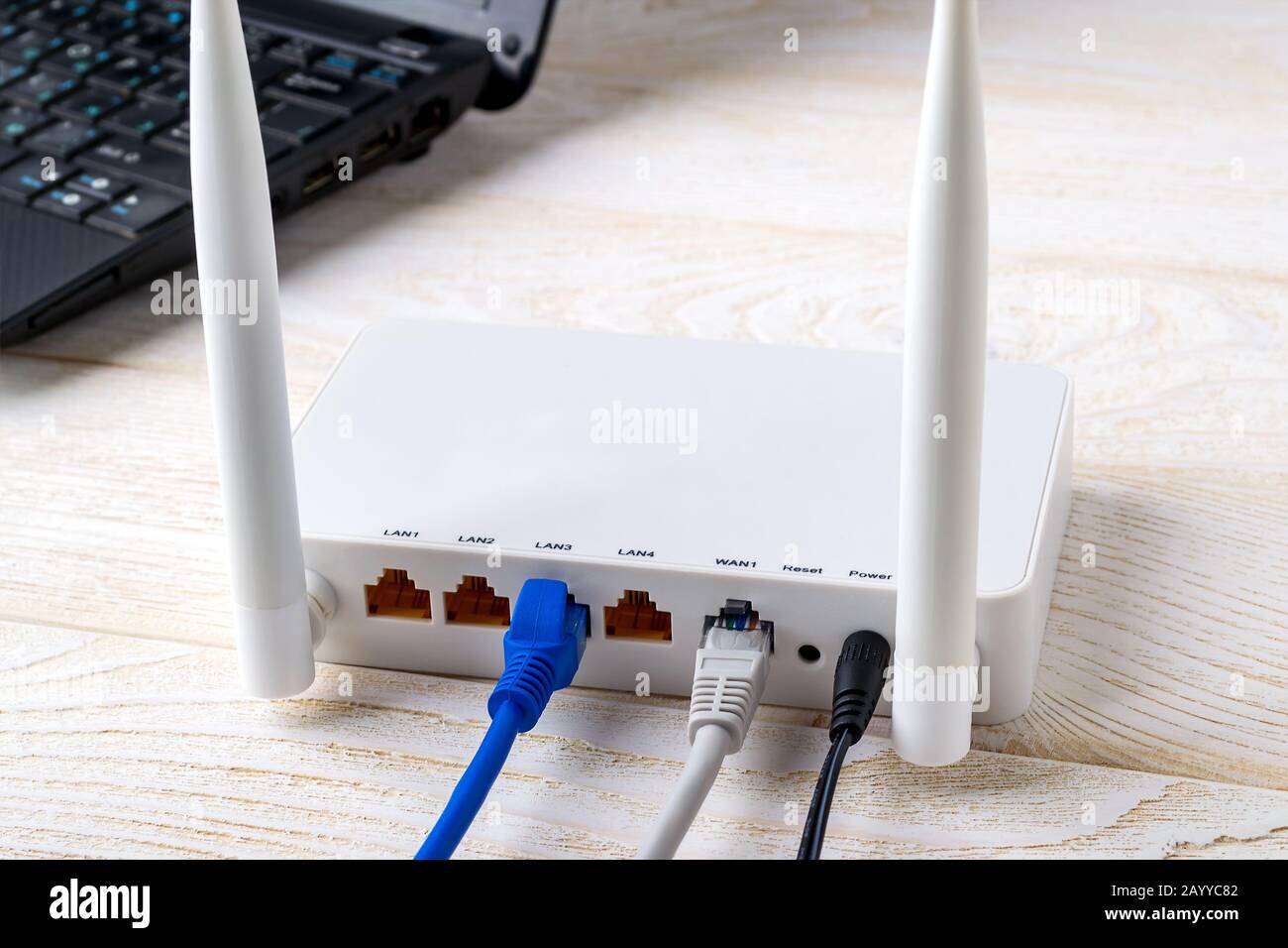 Netzwerkkabel, die mit einem weißen WLAN-Router in der Nähe eines Laptops auf einem weißen Holztisch verbunden sind. WLAN-Router mit auf einem Tisch eingesteckten Internetkabeln Stockfoto