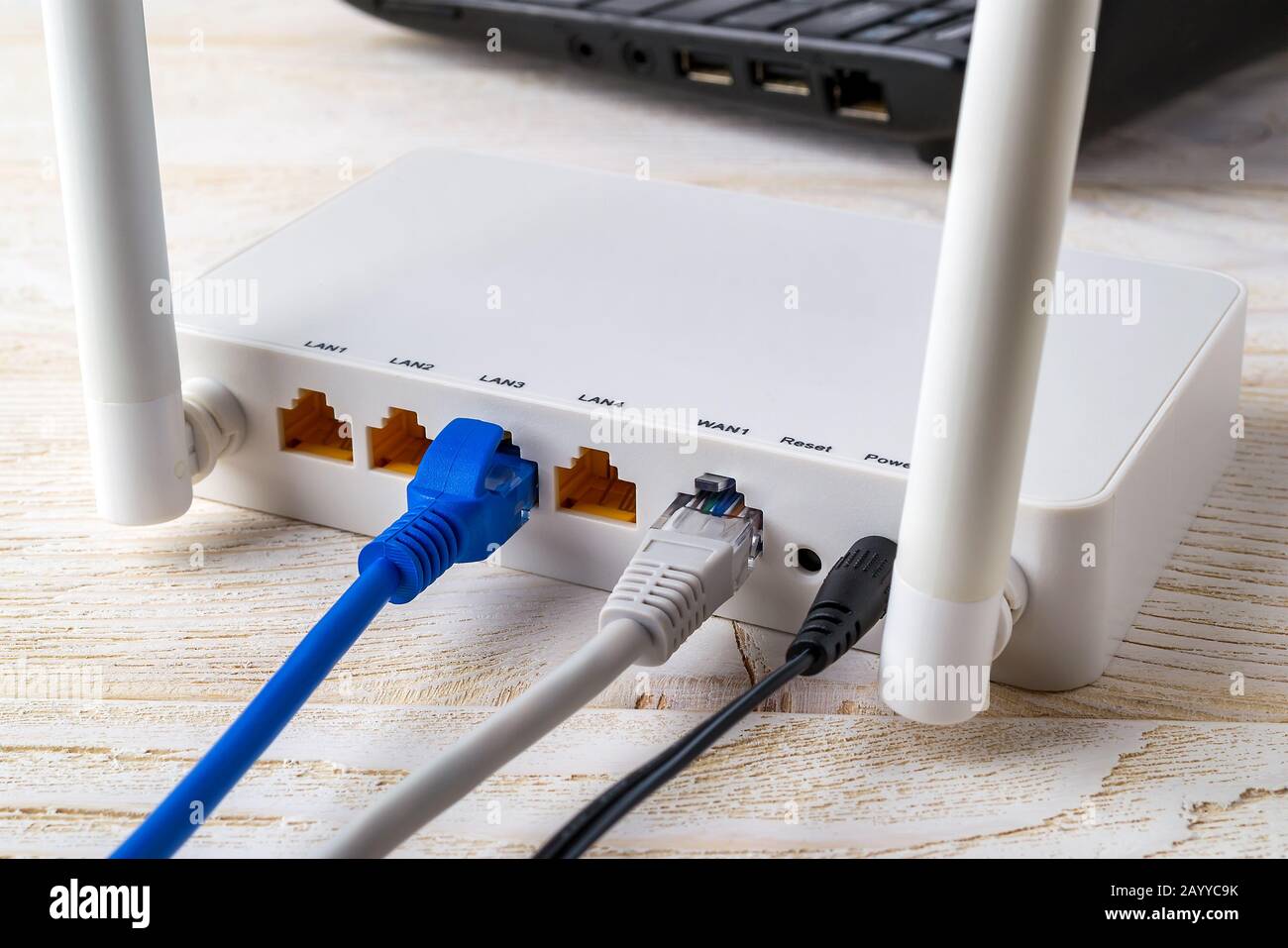 Nahaufnahme der Netzwerkkabel, die mit einem weißen WLAN-Router in der Nähe eines Laptops auf einem weißen Holztisch verbunden sind. WLAN-Router mit eingesteckten Internetkabeln Stockfoto
