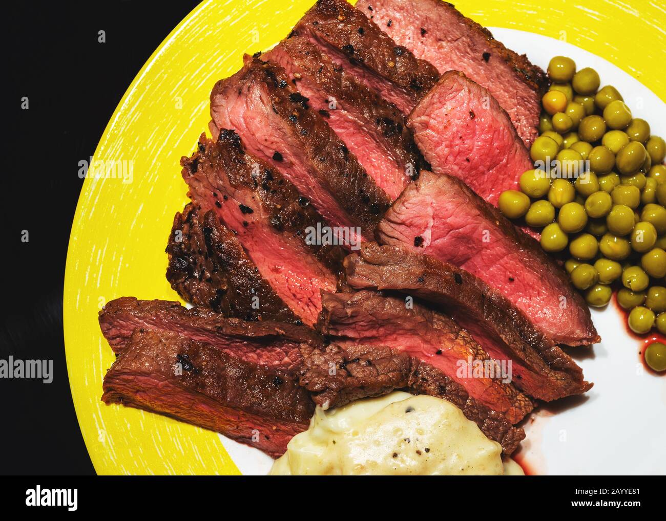 Mittelseltenes hausgemachtes Steak mit Erbsen und Sahnesauce auf gelbem Teller, isoliert auf Schwarz Stockfoto