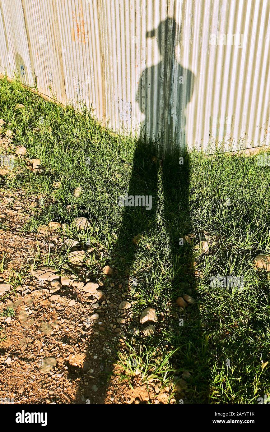 Ein Schatten eines jungen männlichen Menschen mit Hut, nur mit einer Hand grüßenden Gruß, der bei Sonnenuntergang vor einer Wellblechwand und einer Rasenfläche steht Stockfoto