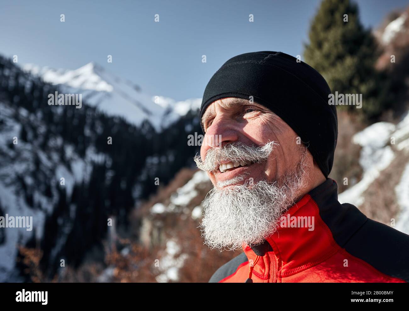 Porträt eines älteren Läufermannes mit grauem Bart, der vor dem Hintergrund der Winterberge lächelt Stockfoto