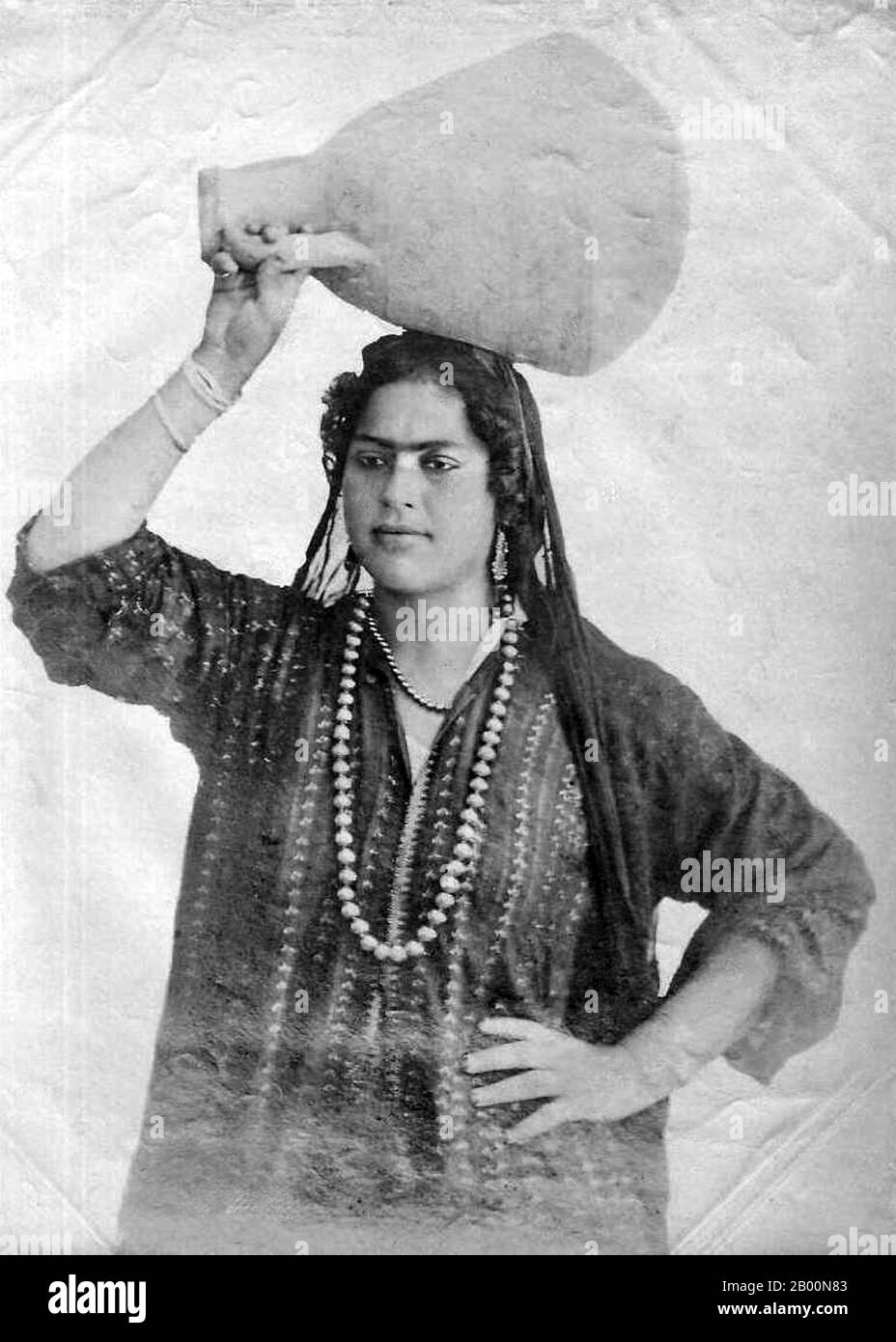Algerien: Porträt einer Basarianin mit Wassertopf auf dem Kopf. Foto einer arabischen Frau aus Algerien, die einen Wassertopf auf dem Kopf trägt. Hergestellt als Souvenir für den entstehenden europäischen Tourismusmarkt. Stockfoto