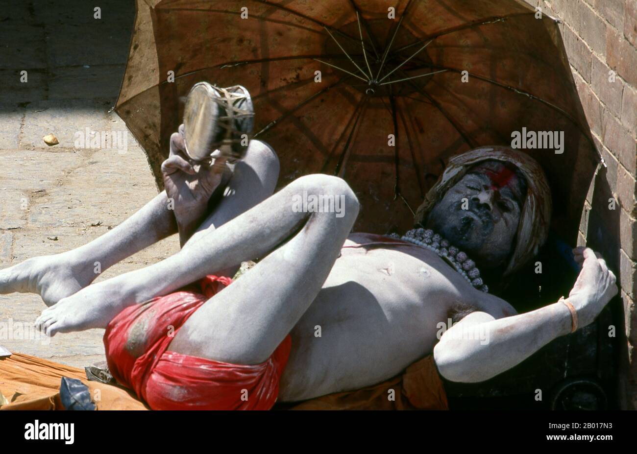 Nepal: Fakir schüttelt sein Tamburin in einer Kathmandu-Straße. Sie sind, verschiedentlich, als Sadhus (Heilige oder 'gute'), Yogis (asketische Praktizierende) bekannt, Fakiren (asketischer Sucher nach der Wahrheit) und Sannyasine (wandernde Bettelmönchen und Asketen). Sie sind die asketischen – und oft exzentrischen – Praktizierenden einer strengen Form des Hinduismus. Geschworen, irdische Wünsche abzuwerfen, entscheiden sich manche dafür, als Anchoriten in der Wildnis zu leben. Andere sind weniger pensioniert, vor allem in den Städten und Tempeln des nepalesischen Kathmandu-Tals. Stockfoto