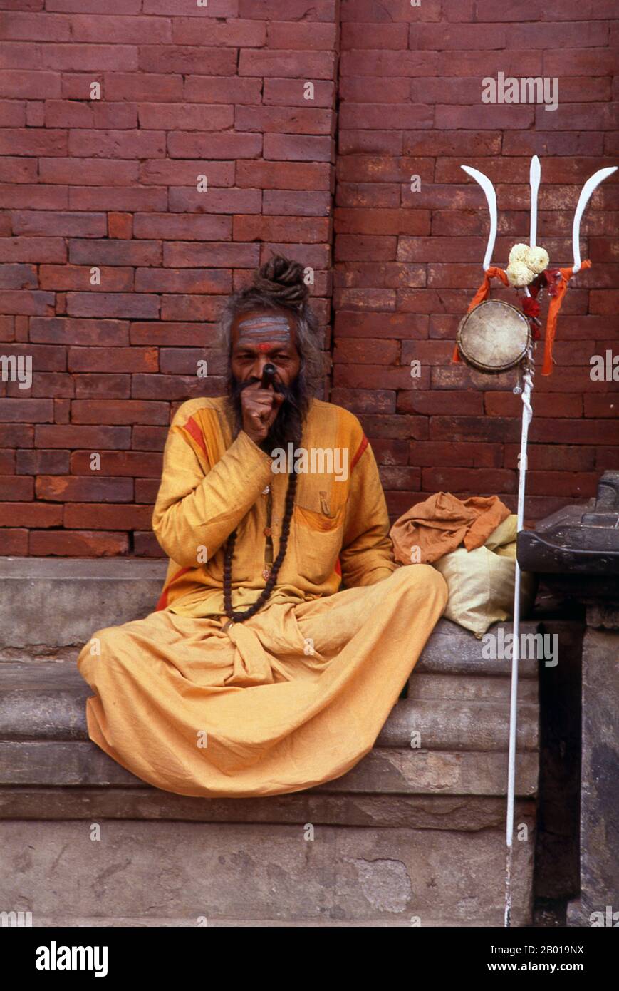 Nepal: Ein Sadhu mit seinem Dreizack und rauchenden Haschisch, Kathmandu. Sie sind, verschiedentlich, als Sadhus (Heilige oder 'gute'), Yogis (asketische Praktizierende) bekannt, Fakiren (asketischer Sucher nach der Wahrheit) und Sannyasine (wandernde Bettelmönchen und Asketen). Sie sind die asketischen – und oft exzentrischen – Praktizierenden einer strengen Form des Hinduismus. Geschworen, irdische Wünsche abzuwerfen, entscheiden sich manche dafür, als Anchoriten in der Wildnis zu leben. Andere sind weniger pensioniert, vor allem in den Städten und Tempeln des nepalesischen Kathmandu-Tals. Stockfoto