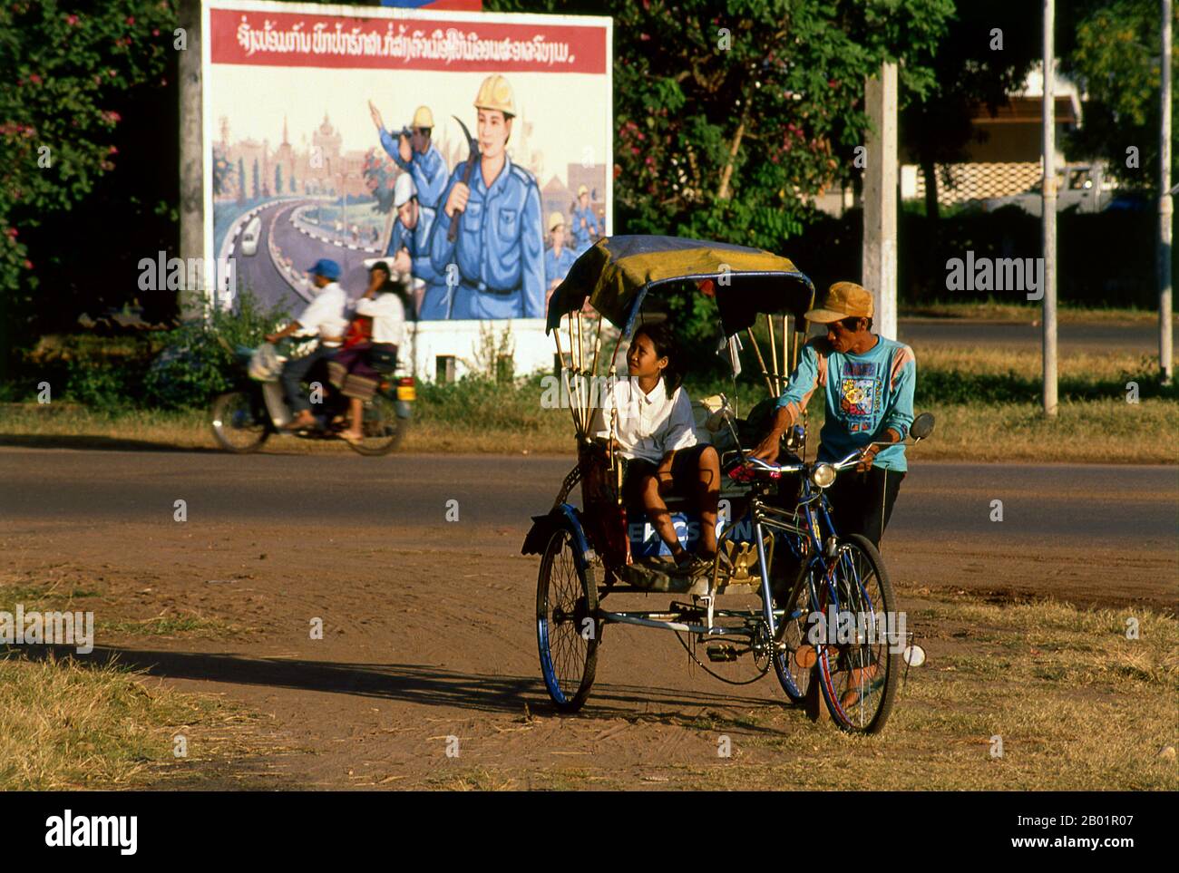 Laos: Ein Pedicab oder Samlo vor einem revolutionären sozialistischen, realistischen politischen Poster auf den Straßen von Vientiane. Der sozialistische Realismus ist ein Stil realistischer Kunst, der in der Sowjetunion entwickelt wurde und in anderen kommunistischen Ländern zu einem dominierenden Stil wurde. Der sozialistische Realismus ist ein teleologisch orientierter Stil, dessen Ziel die Förderung der Ziele des Sozialismus und des Kommunismus ist. Obwohl sie damit verbunden ist, darf sie nicht mit dem sozialen Realismus verwechselt werden, einer Art von Kunst, die Themen sozialer Belange realistisch darstellt. Stockfoto