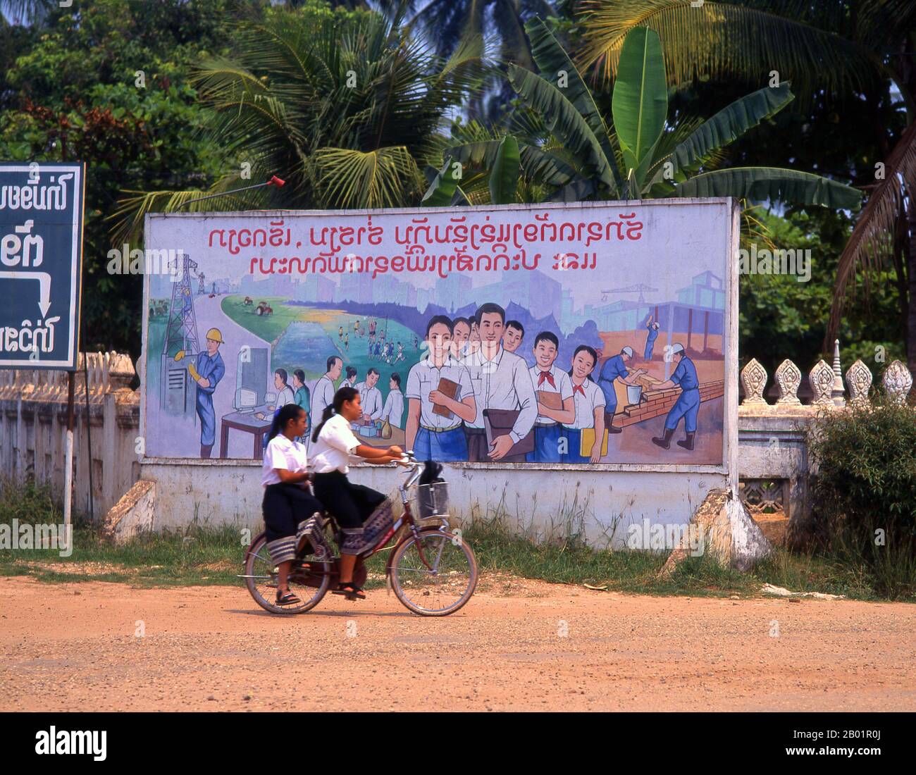 Laos: Zwei Schulmädchen fahren auf den Straßen von Vientiane vor einem sozialistisch-realistischen Bildungs- und Industrieplakat. Der sozialistische Realismus ist ein Stil realistischer Kunst, der in der Sowjetunion entwickelt wurde und in anderen kommunistischen Ländern zu einem dominierenden Stil wurde. Der sozialistische Realismus ist ein teleologisch orientierter Stil, dessen Ziel die Förderung der Ziele des Sozialismus und des Kommunismus ist. Obwohl sie damit verbunden ist, darf sie nicht mit dem sozialen Realismus verwechselt werden, einer Art von Kunst, die Themen sozialer Belange realistisch darstellt. Stockfoto