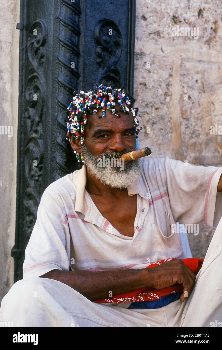 Kuba: Rastaman mit Zigarre auf der Plaza de la Catedral, Old Havanna. Die Plaza de la Catedral ist einer der Plätze der Altstadt von Havanna. Diese gepflasterte, offene Gegend (nur Fußgänger) ist von schönen Gebäuden umgeben und beherbergt die farbenprächtigsten Straßenbewohner und Performancekünstler von La Habana Vieja. Sie reichen von Santería-Priesterinnen über scharfkantige Straßentänzerinnen bis hin zu Blumenmädchen und Rastafariern. Die barocke Catedral de la Habana, erbaut 1777, dominiert den Platz. Offiziell die Catedral de la Virgen María de la Concepción Immaculada, die mit Messing gebundenen Holztüren sind besonders beeindruckend. Stockfoto