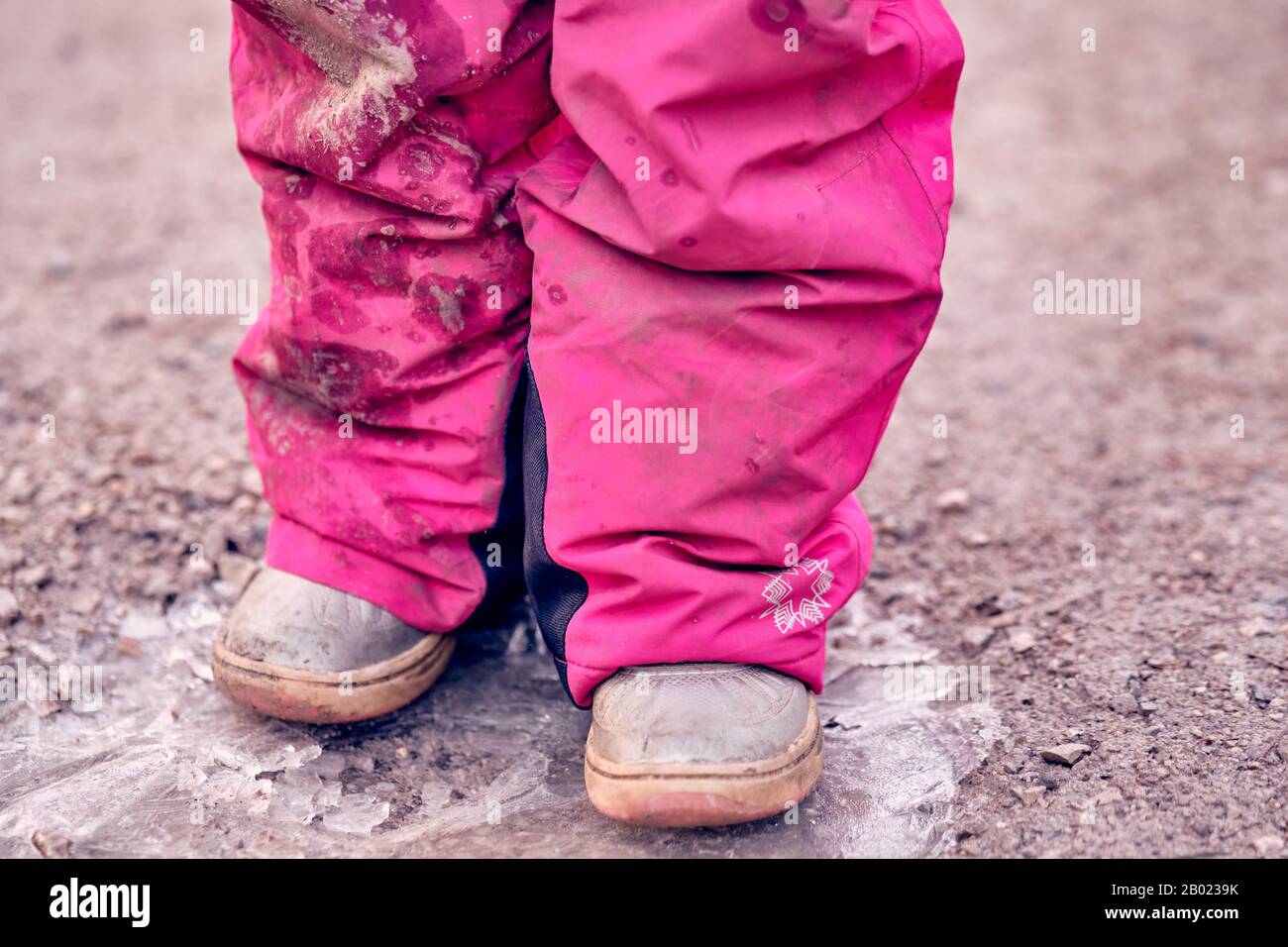 Tiefer Teil eines 4 Jahre alten Kindermädchens in schlammig schmutzig pinkfarbenen Winterhosen, die auf einer gefrorenen Pfütze auf einer Schotterstraße stehen. Im Februar in Deutschland zu sehen. Stockfoto