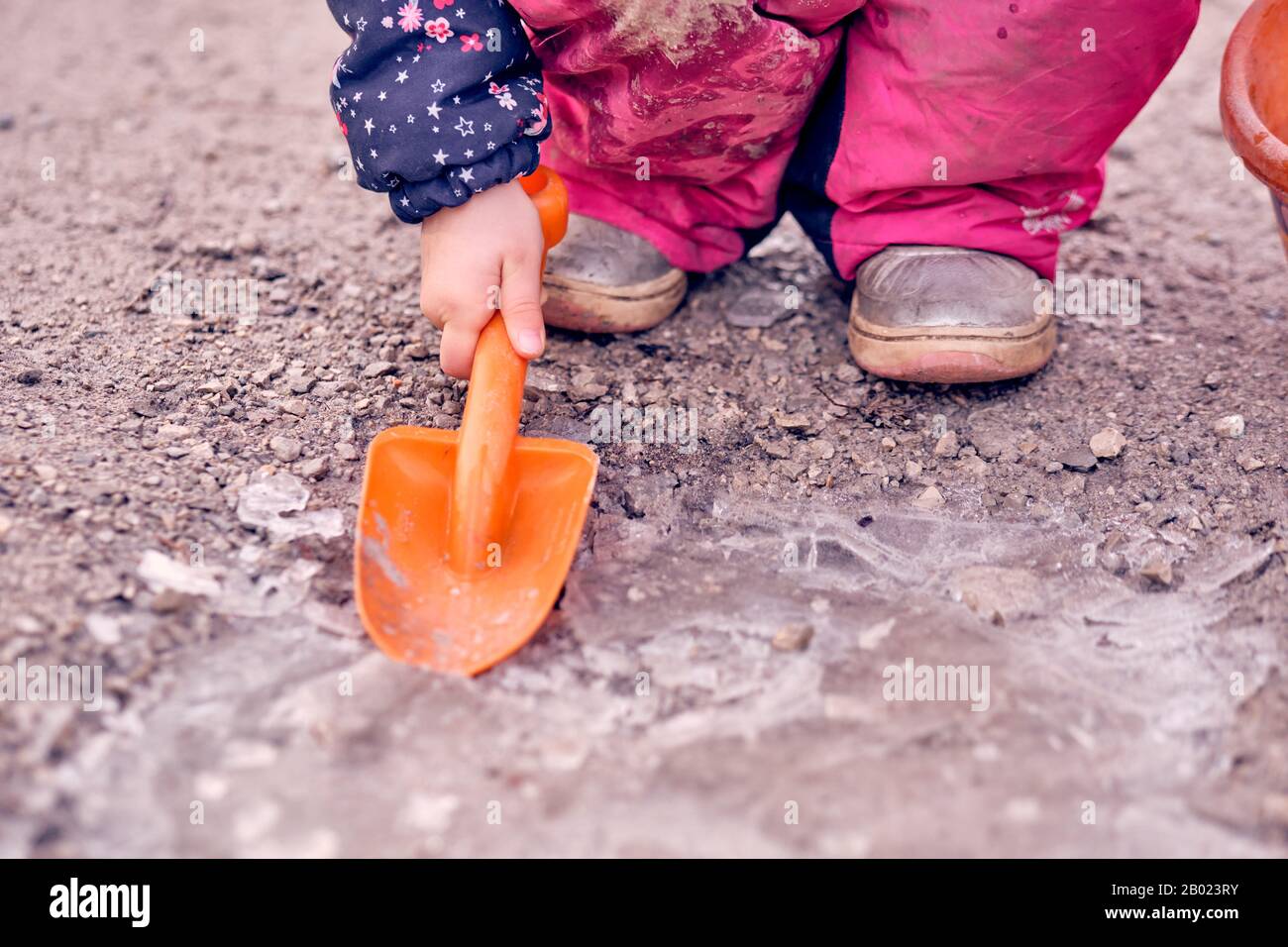 Tiefer Teil eines 4 Jahre alten Kindermädchens in schlammig schmutzig pinkfarbenen Winterhosen, die auf dem Eis an einer gefrorenen Pfütze mit einer orangefarbenen Schaufel hocken und arbeiten. Stockfoto