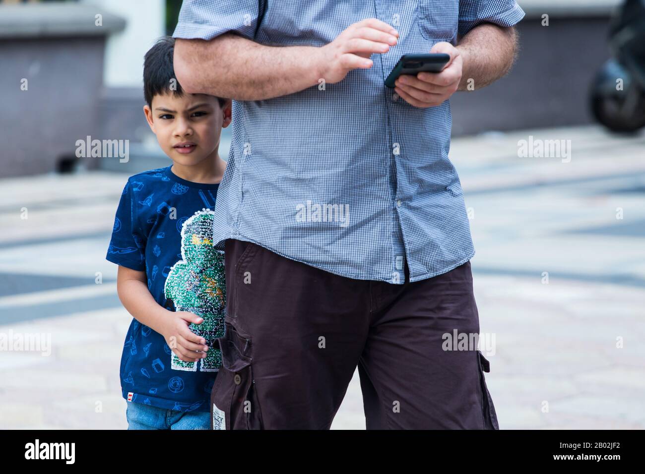 Ein junger, schüchterner arabischer Junge versucht sich hinter seinem Vater zu verstecken, während der Mann oder wahrscheinlich sein Vater mit seinem Handy beschäftigt ist. Stockfoto