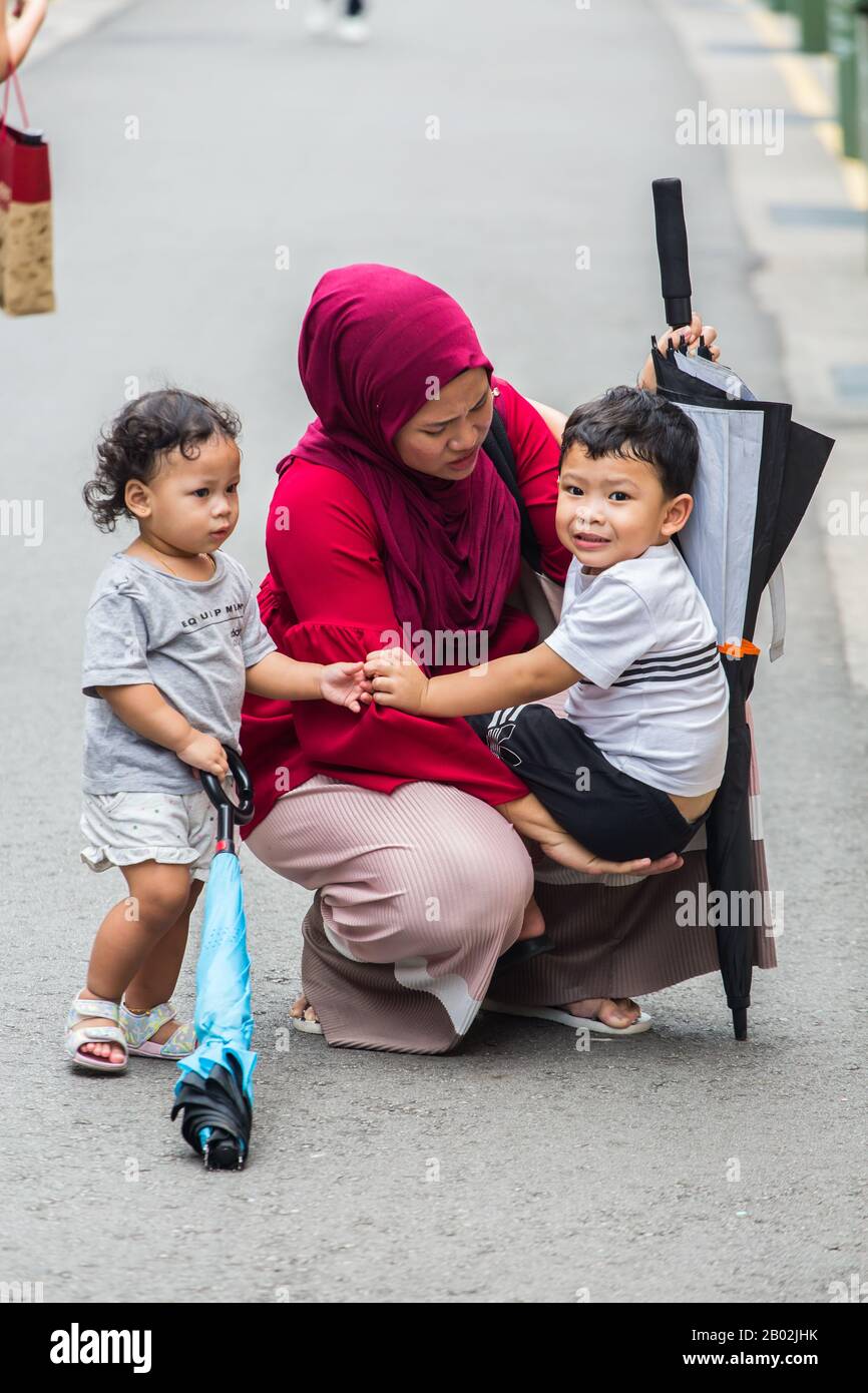 Vertikale Ansicht einer geschäftigen muslimischen Mutter in roten Hijab Squat nach unten zu halten und zu unterstützen ihr weinendes Kind auf der Straße. Singapur. Stockfoto