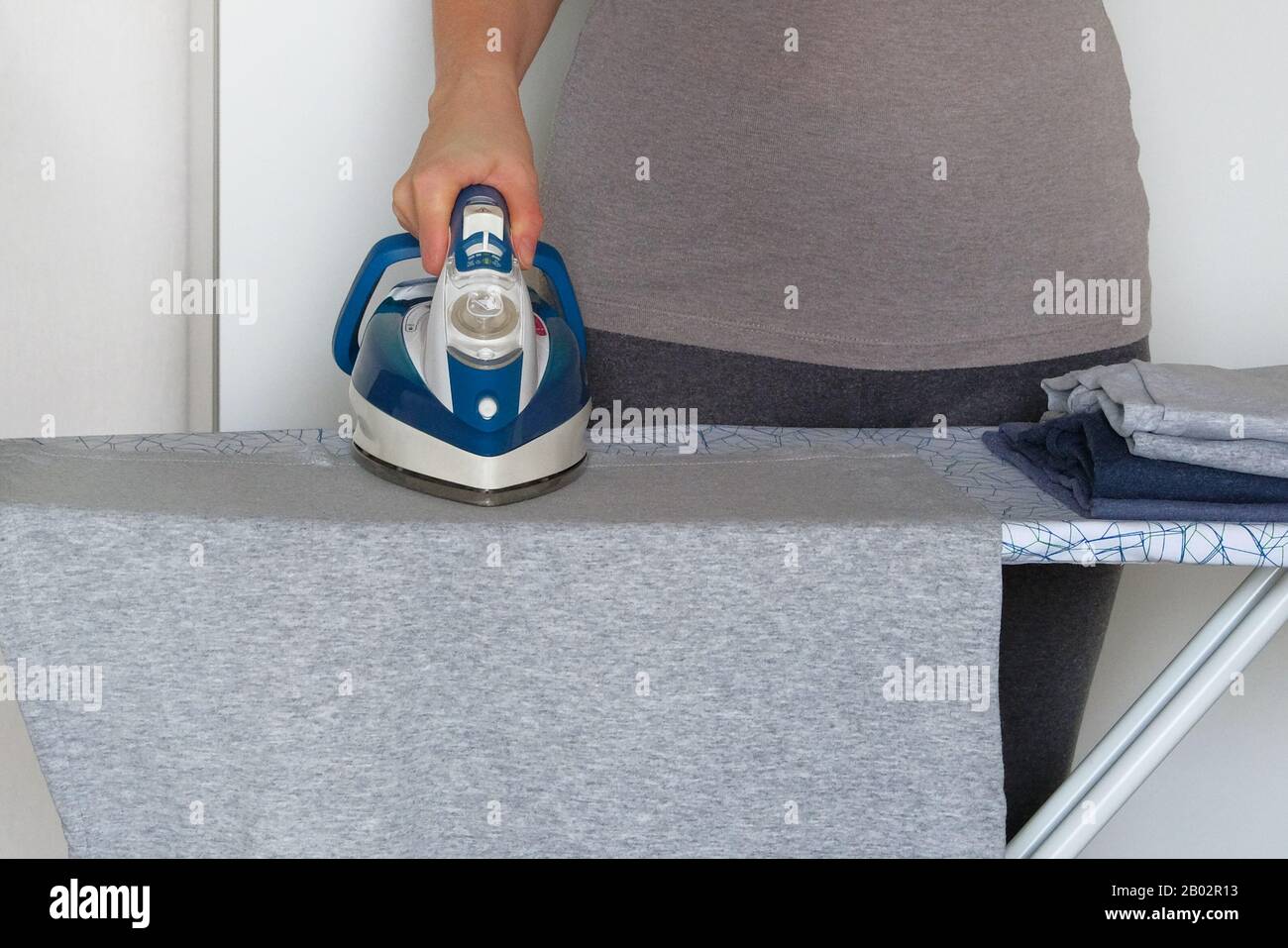 Hausfrau bügelt Kleidung auf dem Bügelbrett mit blauem Bügeleisen. Hausarbeit und Haushaltskonzept. Kleidung bügeln zu Hause. Stockfoto Hausfrau bügelt Kleidung auf dem Bügelbrett mit blauem Bügeleisen. Hausarbeit und Haushaltskonzept. Kleidung bügeln zu Hause. Stockfoto