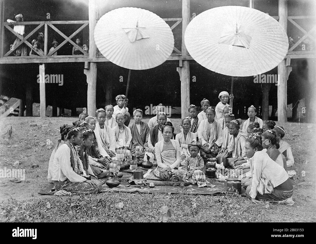 Foto von Sawbwas Frau und Kind in Wuntho in Birma (Myanmar) aus der Sammlung Elgin: "Herbsttour 1898", aufgenommen von Felice Beato c.-2. Das Porträt wurde vor einem Haus in Wuntho aufgenommen und zeigt die Frau und das Kind des Wuntho Sawbwa, eines shan-feudalherren, der mit Lebensmittelkörben auf dem Boden sitzt. Die Teilnehmer sind im Halbkreis um das Paar angeordnet und zwei Diener halten Regenschirme über der Gruppe. Die Frau trägt eine gestreifte Seide hta-mein (Wickelrock), eine eng anliegende Jacke aus feinem Muslin oder Baumwolle, die als ein-Gyi bekannt ist, und Halsketten. Während der Konbaung-Dynastie (1752-1885) wurde am 1. Stockfoto