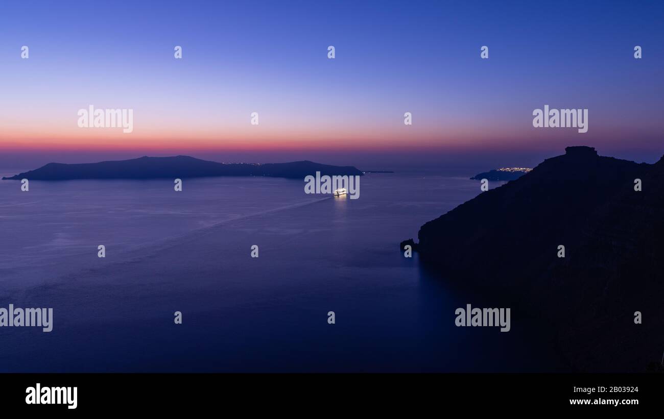 Ein Kreuzfahrtschiff, das kurz nach Sonnenuntergang und während der blauen Stunde aus der Bucht auf der Insel Santorini, Griechenland segelt. Stockfoto