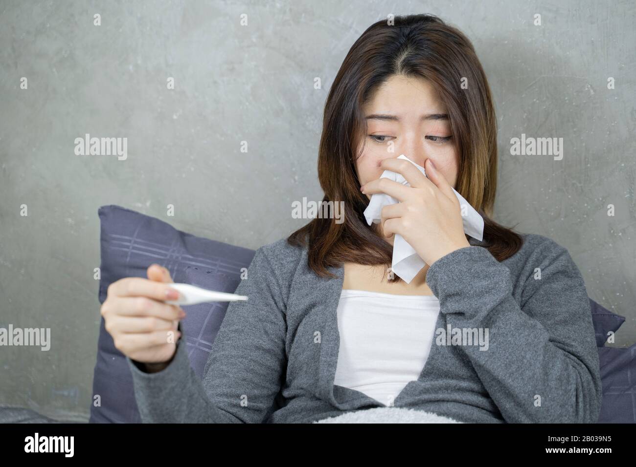 Krankheit spüren und in Papierwisch niesen. Wunderschönes, ungesundes Mädchen, das mit einer Decke bedeckt ist, die Nase wischt und ein Thermometer betrachtet. Konzept für das Gesundheitswesen. Stockfoto