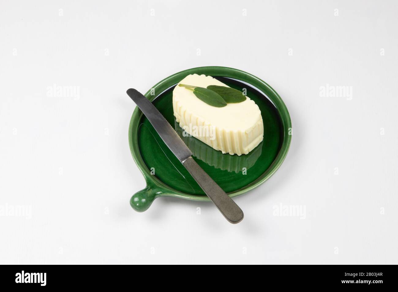 Elegant servierte Butterwürfel auf Keramikschale mit Messer und grünen Salbeiblättern auf weißem Grund. Stockfoto