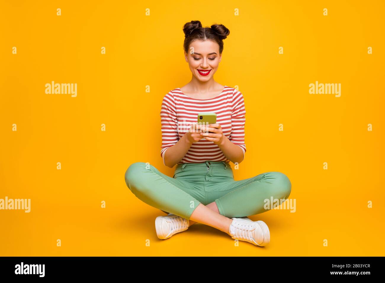 Lange Foto der hübschen attraktiven Dame Sit floor Beine gekreuzt halten Telefon eingeben neuer Post Blog Text tragen gestreiftes rot-weißes Hemd grüne Hosen Stockfoto