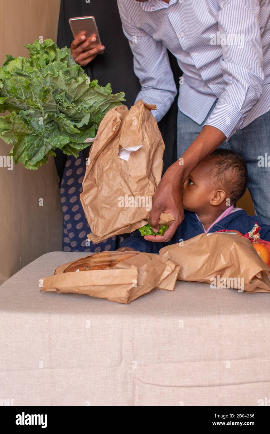 Unerkennbarer Afrikaner packt Gemüse mit seiner Familie aus, als ein Baby neugierig ihre Art drängt, zu versuchen, an die Kale zu kommen, die der Mann hält Stockfoto