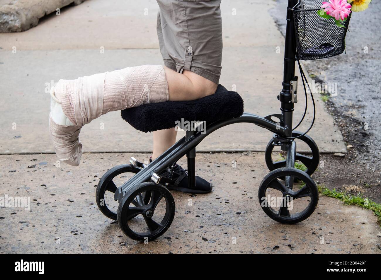 Frau mit gewickeltem Bein auf lenkbarem Kniehauser Roller mit Blumen auf Korb und Handy innen auf rauer zerbrochener Zementoberfläche - beschnitten Stockfoto