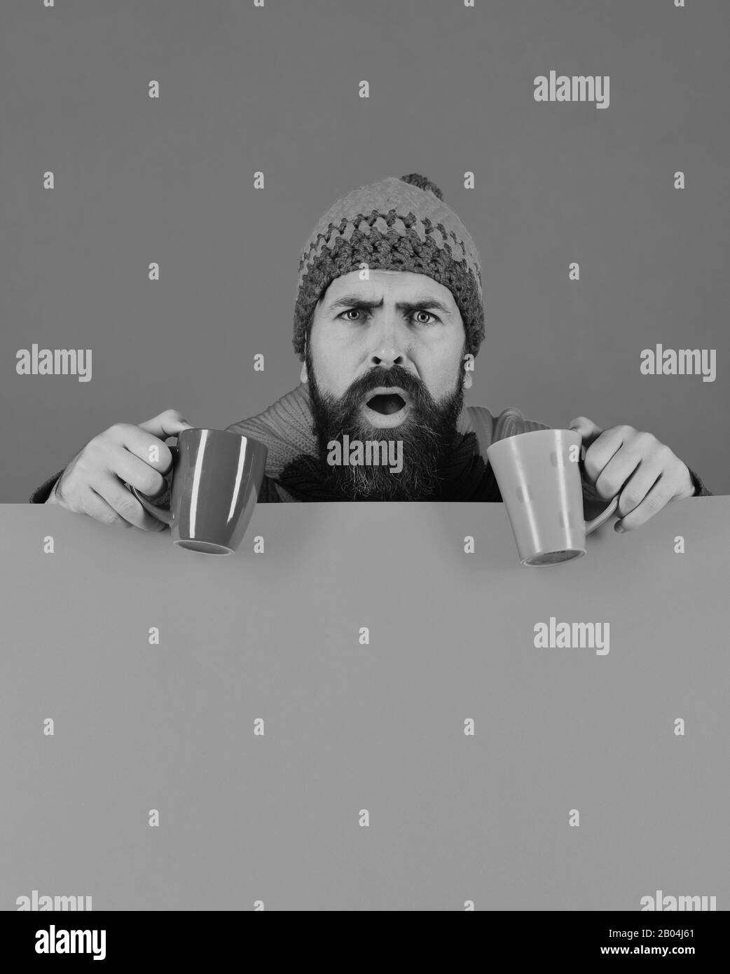 Mann in warme Mütze hält braune und grüne Tassen auf grün und orange Hintergrund, kopieren. Herbst und heiße Getränke Konzept. Getränke und gemütliche Zeit. Hipster mit Bart und entsetzte Gesicht hat warmen Tee Stockfoto