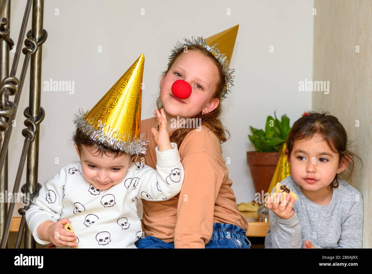 Portrait von Teenager-Mädchen mit einer Clownnase, die sich mit kleinen Kindern in einem Partyhut amüsieren, der Plätzchen auf dem Purim oder der Geburtstagsfeier isst. Stockfoto