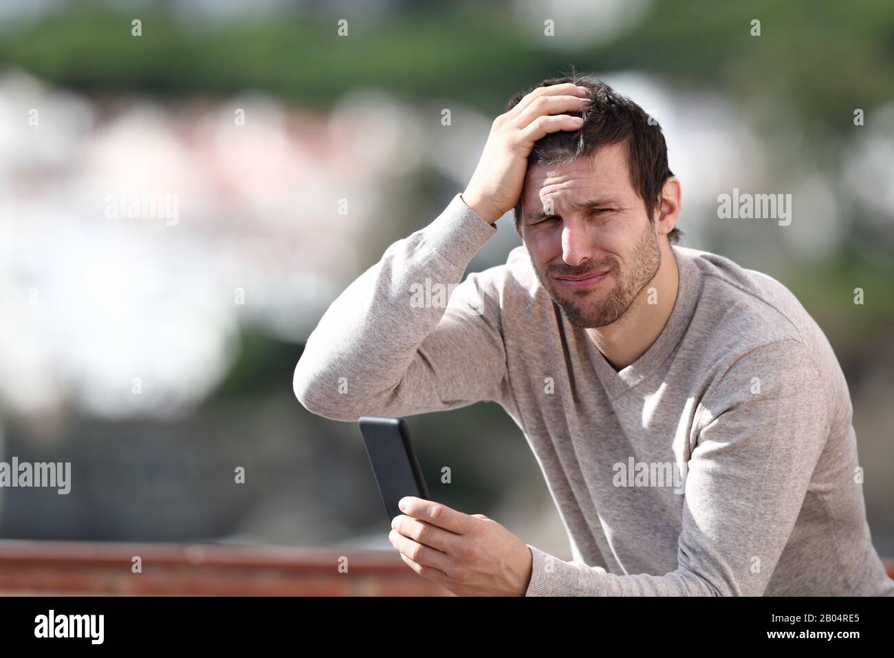 Verwirrter Mann, der sein Handy hält, beschwert sich nach einem Fehler, in einer ländlichen Stadt zu sitzen Stockfoto