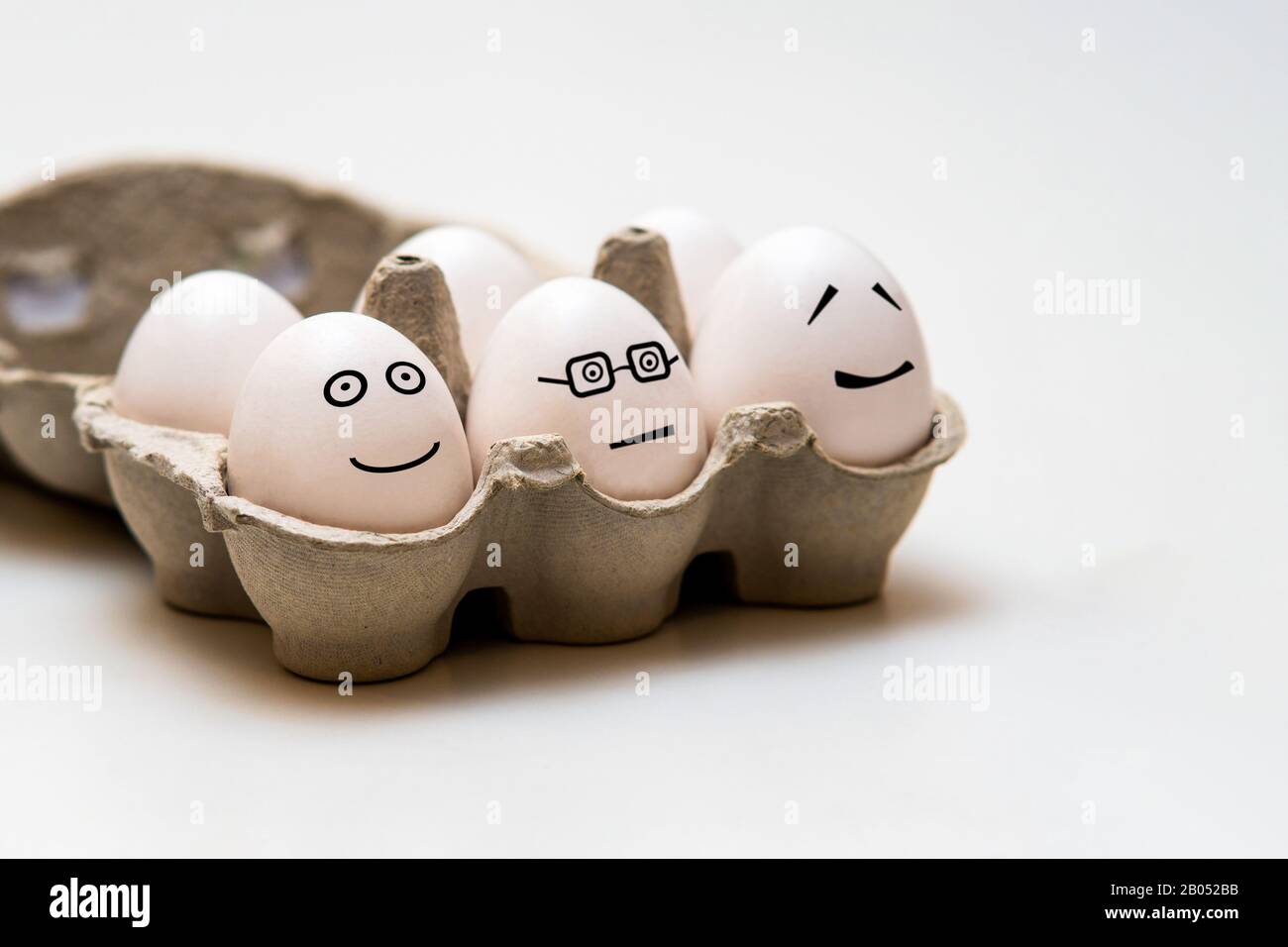Weiße Eier mit lustigen Gesichtern in der braunen kleinen Aufbewahrungsbox auf hellem Hintergrund Stockfoto
