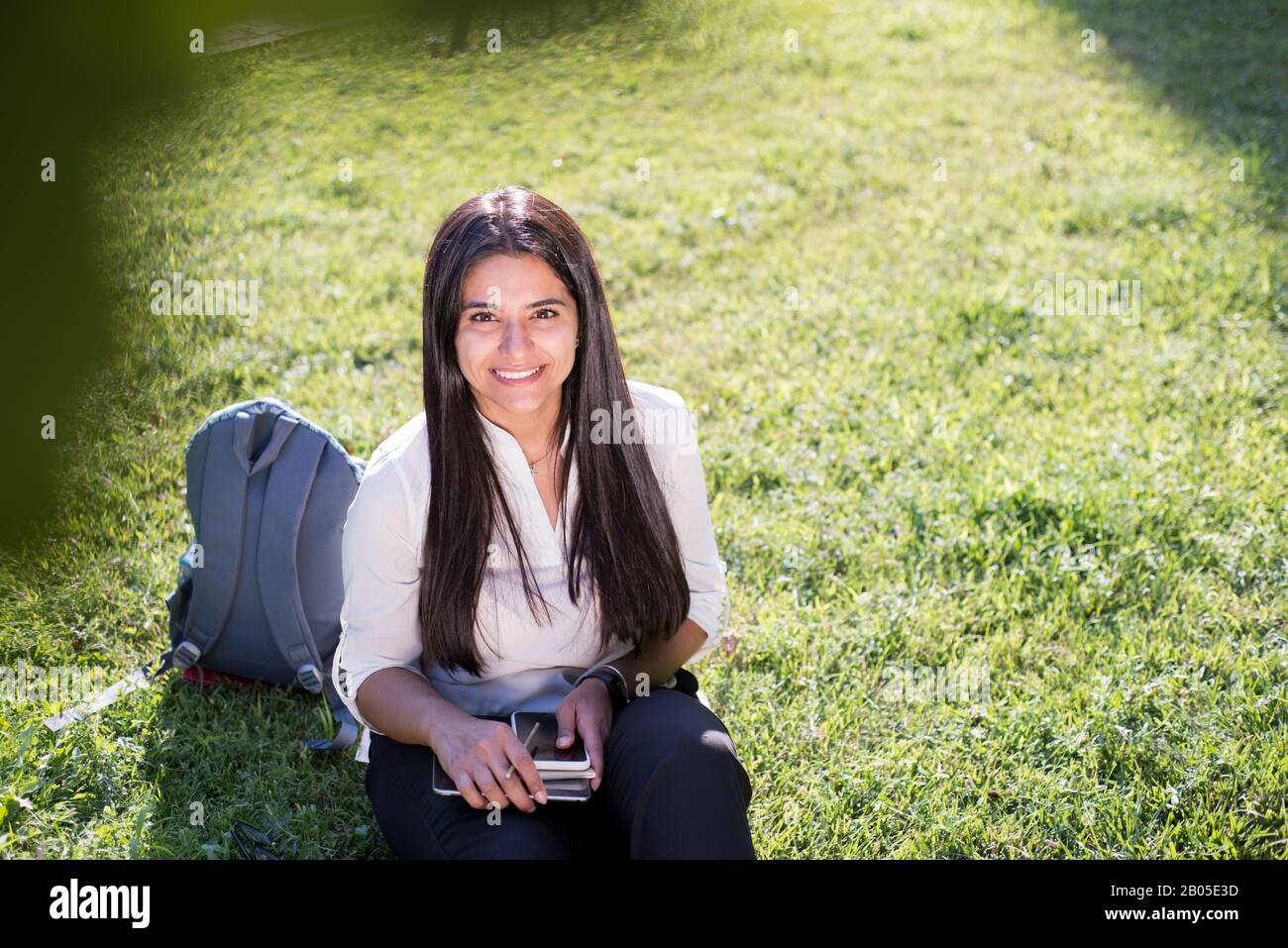 Porträt eines jungen indischen Studenten, Geschäftsfrau. Eine Frau mit einem Rucksack in einem Rucksack hält eine Tablette, sitzt auf dem grünen Gras, gegen die Sonne. Su Stockfoto