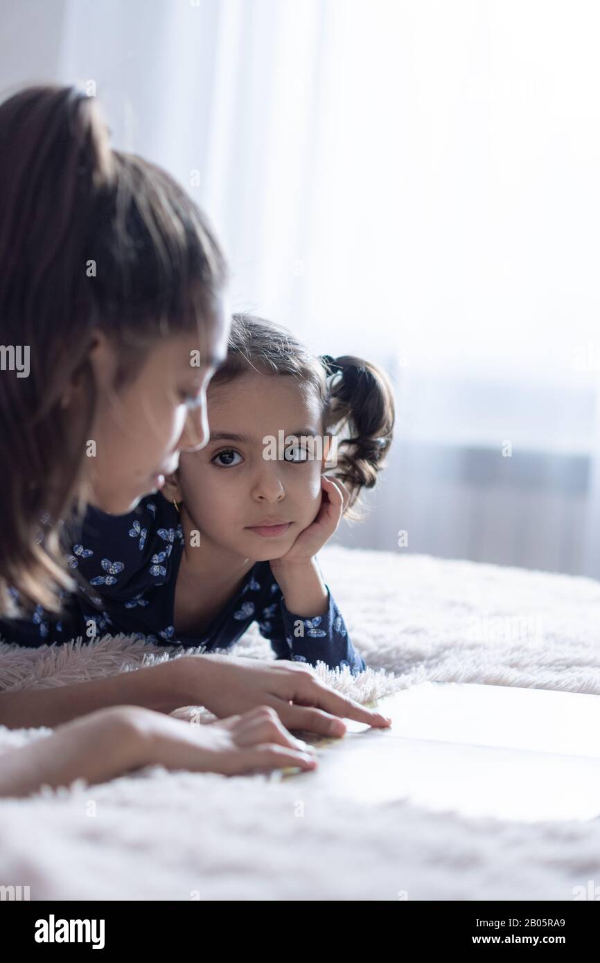 Ein kleines Mädchen mit schwarzen Augen sieht ihre Schwester an, die einen Finger auf ein Buch zeigt. Ein dunkelhäutiges Baby aus dem Nahen Osten sieht ihre Schwester an Stockfoto