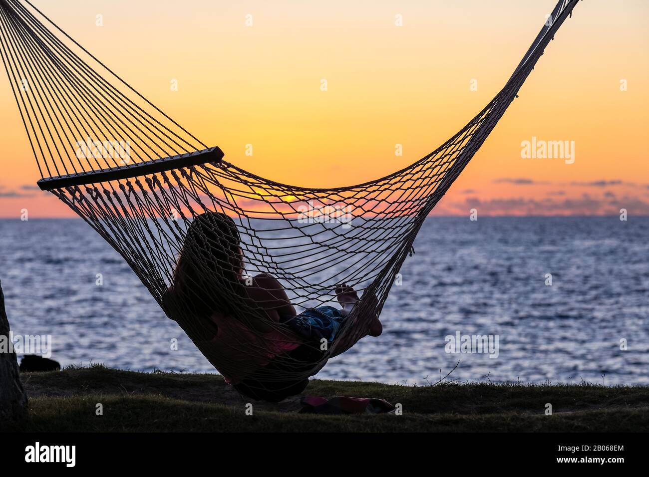 Silhouette einer jungen Frau, die in einer Hängematte ruht und den Sonnenuntergang über dem Pazifischen Ozean, Big Island, Hawaii, beobachtet Stockfoto