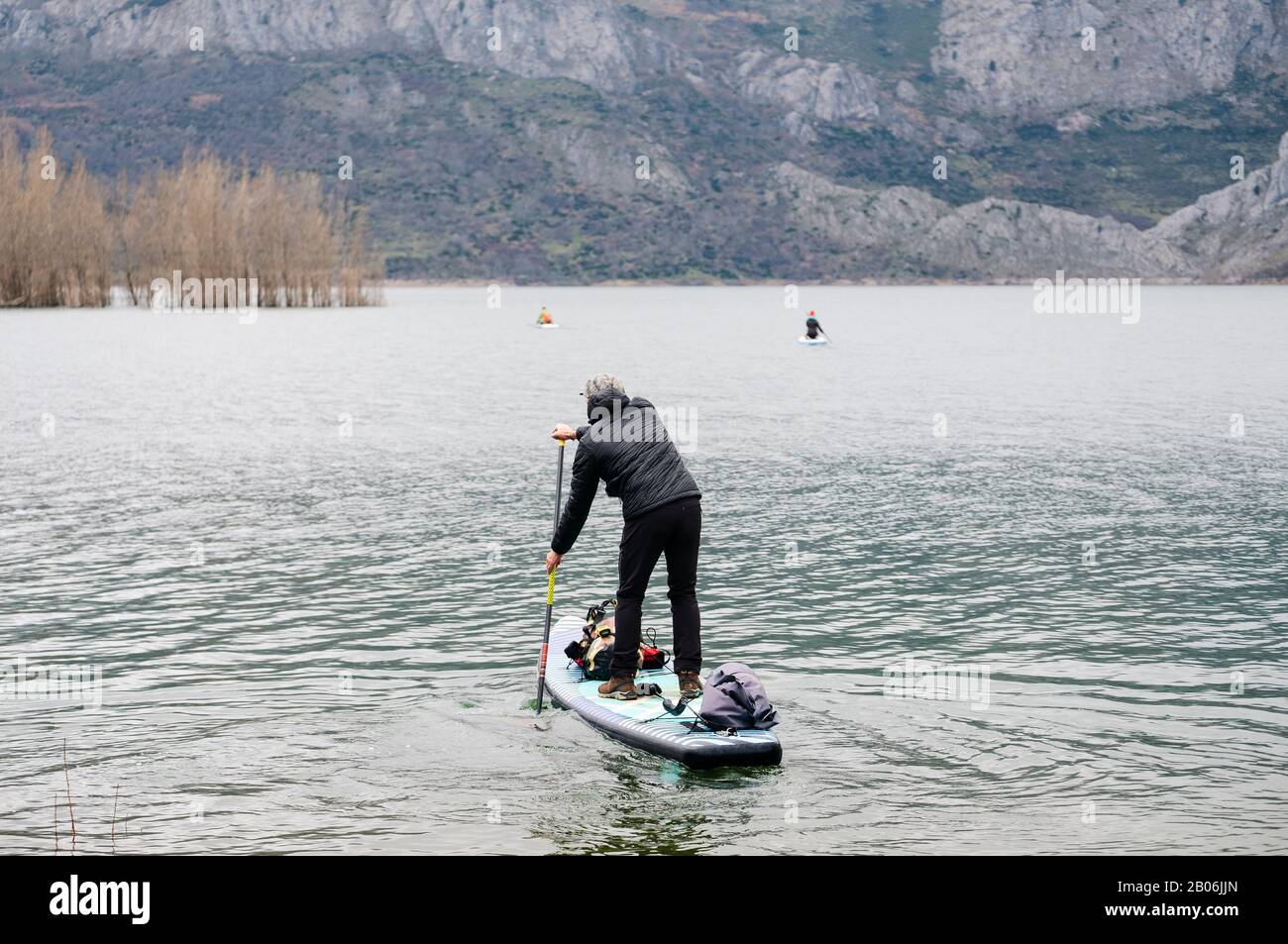 Ein Mann mit warmer Kleidung macht das Surfen im Paddel in einem Bergsee mit Gepäck auf seinem Brett zu erkunden Stockfoto