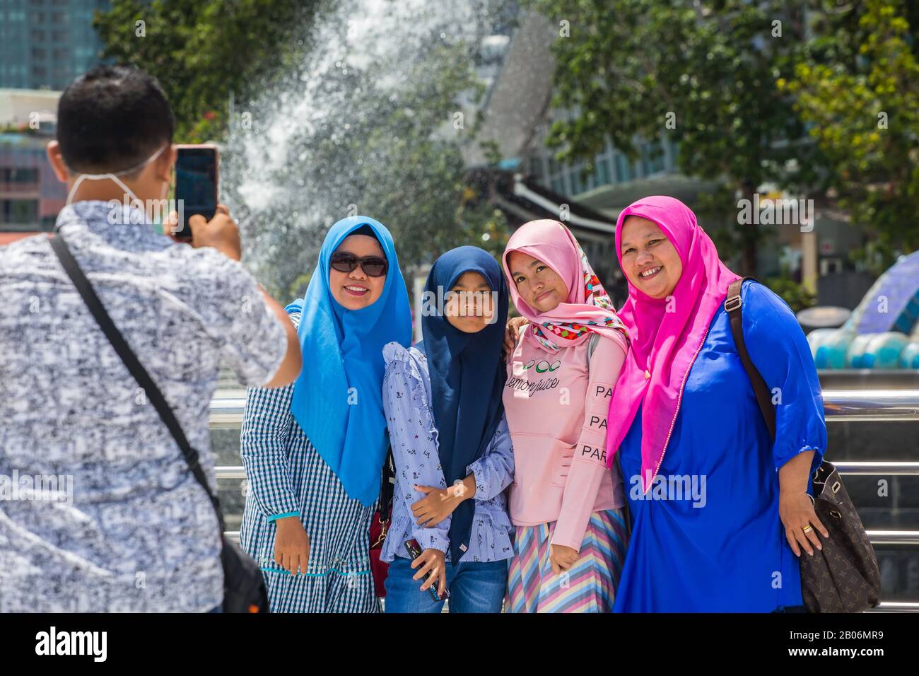 Ein Mann, der mit einem Smartphone fotografiert, um eine Gruppe muslimischer indonesischer Damen zu fotografieren, die natürlich im Merlion Park in Singapur posieren. Stockfoto