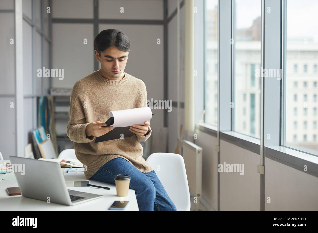 Ein ernsthafter junger Geschäftsmann in legerer Kleidung, der an seinem Arbeitsplatz sitzt und ein Dokument im Büro liest Stockfoto