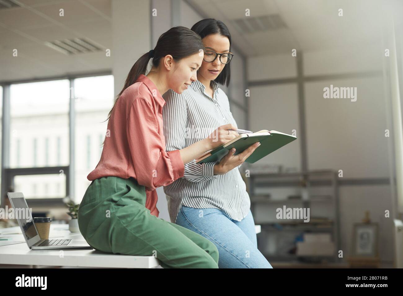 Zwei junge Geschäftsfrauen in legerer Kleidung mit Notizblock und Blick auf ihre Pläne für eine Arbeitswoche im Büro Stockfoto