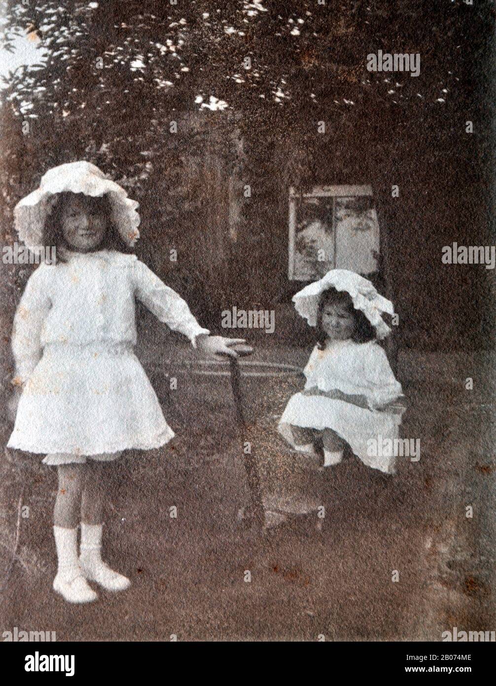 Archivfoto, alt, schwarz und weiß von zwei jungen Mädchen in einem Garten, von denen eines in einem Rollwagen sitzt, während das ältere Mädchen den Griff des Rollwagens hält, als ob sie ihn zieht. Beide Mädchen tragen weiße Kleider und weiße Hüte. Stockfoto