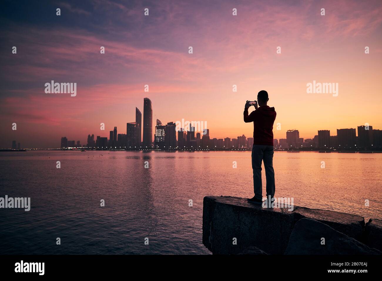 Junger Mann mit Handy fotografiert bei Sonnenaufgang die Skyline der Stadt. Abu Dhabi, Vereinigte Arabische Emirate. Stockfoto